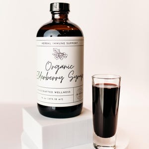 Könnte beinhalten: Eine Glasflasche mit Bio-Holunderbeersaft mit einem weißen Etikett, auf dem "Herbal Immune Support" und "Organic Elderberry Syrup" steht. Die Flasche steht auf einer weißen Plattform neben einem Glas, das mit dem dunkelvioletten Sirup gefüllt ist.
