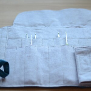 May include: A light gray linen crochet hook organizer, open to display several crochet hooks. The organizer has multiple pockets for the hooks and a teal strap. The design is simple and functional.