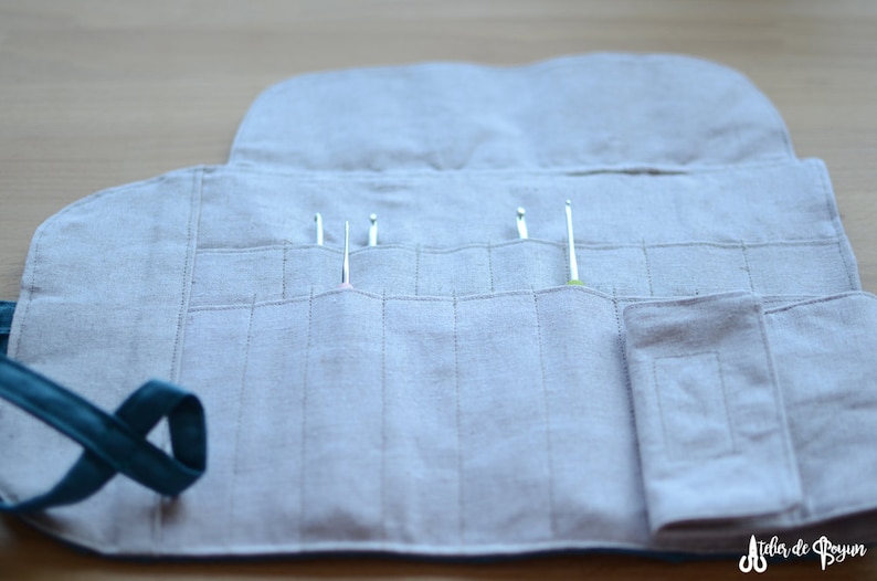 May include: A light gray linen crochet hook organizer, open to display several crochet hooks. The organizer has multiple pockets for the hooks and a teal strap. The design is simple and functional.
