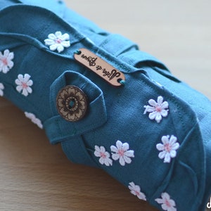 May include: Teal fabric roll-up case adorned with white daisy appliqu&eacute;s with pink centers. Features a wooden button and a small leather tag with text. The case is designed with leaf-shaped accents.