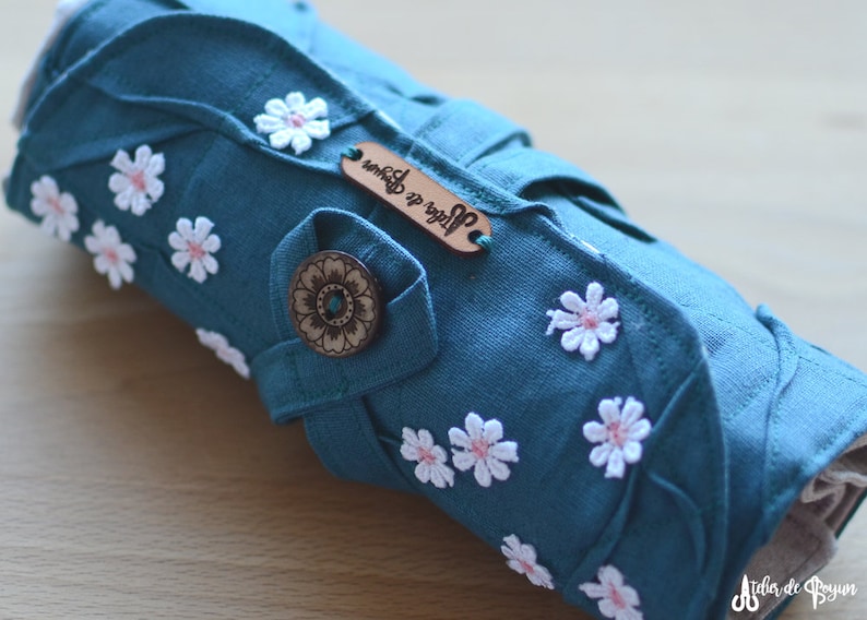 May include: Teal fabric roll-up case adorned with white daisy appliqu&eacute;s with pink centers. Features a wooden button and a small leather tag with text. The case is designed with leaf-shaped accents.