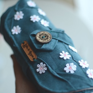 May include: Teal fabric case adorned with white daisy appliqu&eacute;s and a wooden button closure. A small leather tag with the text "Atelier de Boyun" is attached. The case is designed with a folded flap and decorative stitching.