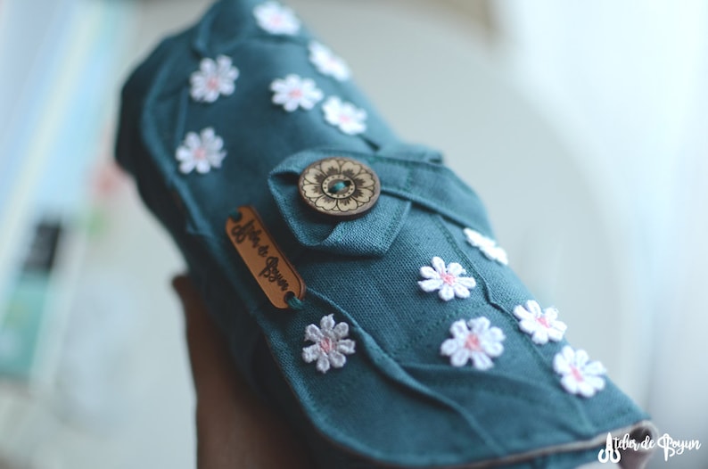 May include: Teal fabric case adorned with white daisy appliqu&eacute;s and a wooden button closure. A small leather tag with the text "Atelier de Boyun" is attached. The case is designed with a folded flap and decorative stitching.