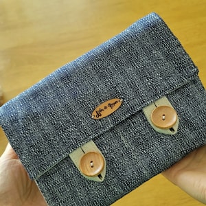 May include: A denim-style clutch with a flap closure, secured by two wooden buttons. The clutch features a small leather tag with the text "Affinity & Bloom". The fabric has a speckled pattern.