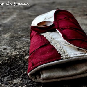 May include: A handmade, folded wallet with a button closure. The wallet features a patchwork design with burgundy and cream-colored fabric, accented with lace trim. The text "Atelier de Soyun" is visible in the upper left corner.