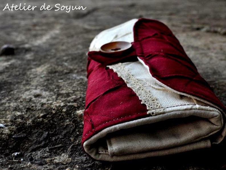 May include: A handmade, folded wallet with a button closure. The wallet features a patchwork design with burgundy and cream-colored fabric, accented with lace trim. The text "Atelier de Soyun" is visible in the upper left corner.