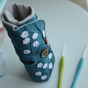 May include: Teal fabric crochet hook case with white daisy appliqu&eacute;s and a wooden button closure. The case is open, revealing a gray interior lining. Two crochet hooks, one green and one blue, are next to the case.