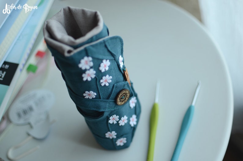 May include: Teal fabric crochet hook case with white daisy appliqu&eacute;s and a wooden button closure. The case is open, revealing a gray interior lining. Two crochet hooks, one green and one blue, are next to the case.