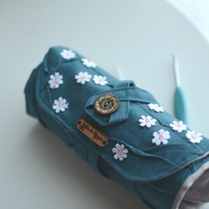 May include: Teal fabric crochet hook case with white daisy embellishments and a wooden button closure. The case has a leather tag with text. A teal crochet hook is visible in the background. The case is rolled up.