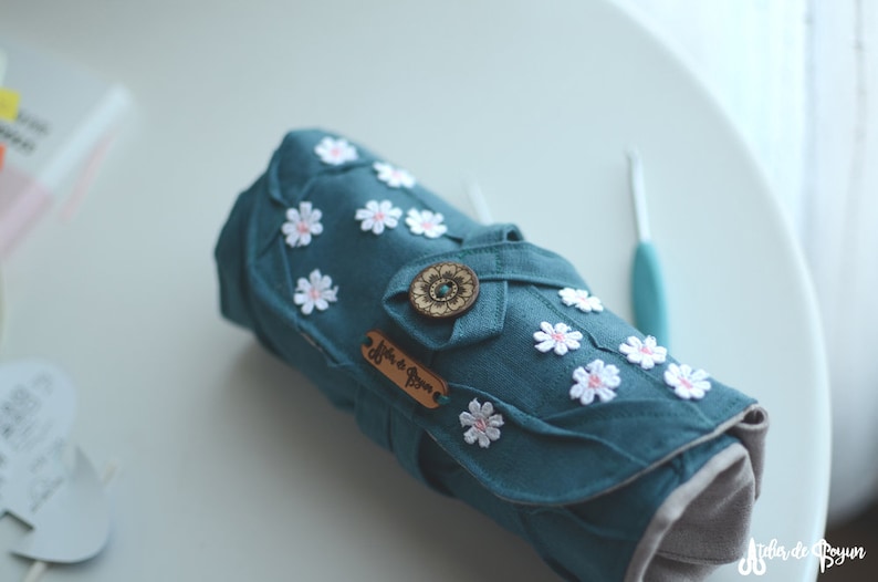 May include: Teal fabric crochet hook case with white daisy embellishments and a wooden button closure. The case has a leather tag with text. A teal crochet hook is visible in the background. The case is rolled up.