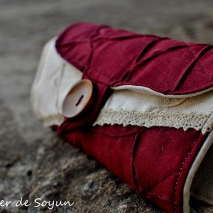 May include: A burgundy and cream-colored fabric clutch with a button closure. The clutch features a quilted design on the burgundy fabric and a cream-colored flap with lace trim. The text "Atelier de Soyun" is visible.