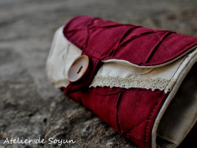 May include: A burgundy and cream-colored fabric clutch with a button closure. The clutch features a quilted design on the burgundy fabric and a cream-colored flap with lace trim. The text "Atelier de Soyun" is visible.