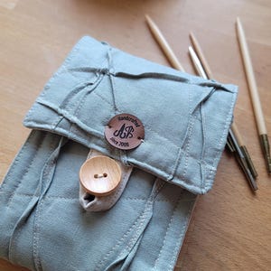 May include: A sage green fabric case with a wooden button closure and a circular brown leather tag featuring the text Handcrafted AB since 2008. The case is accompanied by a ball of light pink yarn and several wooden knitting needles.