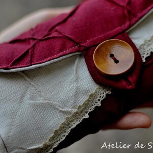 May include: A handmade, burgundy and cream-colored fabric wallet with a wooden button closure. The wallet features a quilted burgundy flap and a cream-colored body with delicate lace trim. The Atelier de Soyun logo is visible in the bottom right corner.