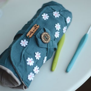 May include: A teal crochet hook case adorned with white daisy appliqu&eacute;s and a wooden button closure. The case is rolled up, with two crochet hooks in green and blue lying beside it. The case has the text "Atelier de Boyun" on a wooden tag.