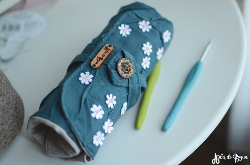 May include: A teal crochet hook case adorned with white daisy appliqu&eacute;s and a wooden button closure. The case is rolled up, with two crochet hooks in green and blue lying beside it. The case has the text "Atelier de Boyun" on a wooden tag.