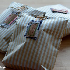 May include: Three wrapped packages with brown and white striped paper. Each package has a fabric tag attached with twine. The tags have decorative stitching and a vintage aesthetic. The packages are arranged on a wooden surface.