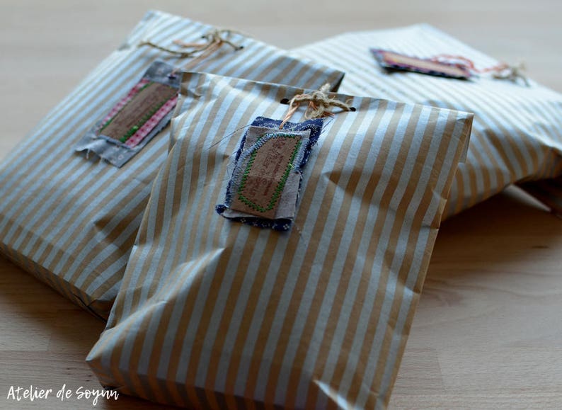 May include: Three wrapped packages with brown and white striped paper. Each package has a fabric tag attached with twine. The tags have decorative stitching and a vintage aesthetic. The packages are arranged on a wooden surface.