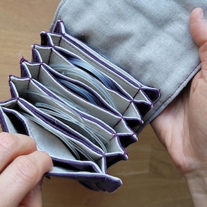 May include: A gray fabric knitting needle case with purple trim and multiple accordion-style pockets. The case is open, revealing several clear, flexible knitting needles. The case is held open by a person's hands.