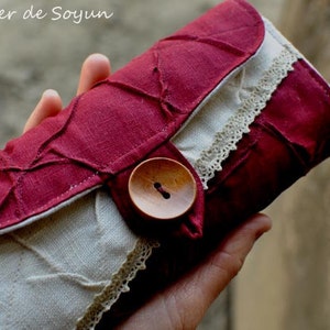 May include: A handmade, burgundy-colored fabric clutch with a decorative quilted design. The clutch features a beige lace trim and a wooden button closure. The text "Atelier de Soyun" is visible at the top.