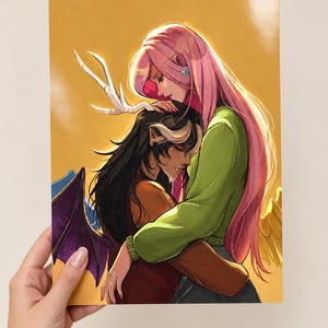 May include: A rectangular art print featuring a digital illustration of two figures embracing against a solid yellow background. One figure has long pink hair and a green sweater, while the other has dark hair, white antlers, and purple wings.