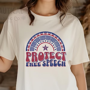 May include: Ivory-coloured t-shirt with a rainbow design, stars, and the words "PROTECT FREE SPEECH" in red and blue. The shirt has short sleeves and a crew neck. The text "IVORY" is in a pink circle.