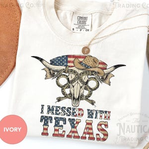 Może przedstawiać: Kremowa koszulka z grafiką przedstawiającą czaszkę byka, kapelusz kowbojski i flagę amerykańską. Tekst "I Messed With Texas" jest wydrukowany poniżej. Widoczny jest również złoty naszyjnik i brązowy kapelusz. Koszulka jest z 'Comfort Colors'.