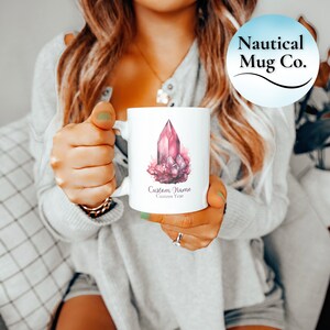 Puede incluir: Una taza de cerámica blanca con una ilustración en acuarela de un grupo de cristales rosas. Debajo del cristal, el texto dice Custom Name y Custom Year en una fuente cursiva. La taza tiene un asa estándar y un acabado blanco brillante.