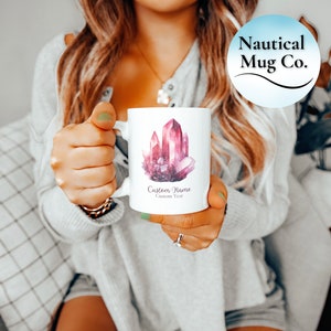 Peut inclure: Un mug en céramique blanche orné d'une illustration style aquarelle de cristaux roses et violets. Sous le motif de cristal, le texte indique Custom Name et Custom Year. Le mug possède une anse standard et une finition lisse et brillante.