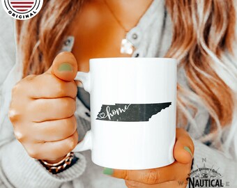 Puede incluir: Taza de café de cerámica blanca con un contorno negro del estado de Tennessee y la palabra "home" escrita en cursiva con un corazón delante de la palabra. La taza está siendo sostenida por la mano de una persona.