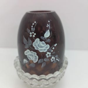 May include: A dark brown, oval-shaped glass vase with a clear, scalloped base. The vase is adorned with hand-painted white roses and delicate floral accents. This decorative vase is suitable for displaying flowers or as a standalone decorative piece.