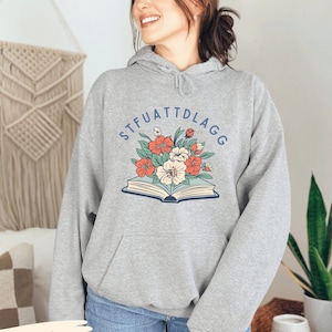 May include: A gray hooded sweatshirt with the text "STFUATTDLAGG" printed in blue on the front. The text is surrounded by a floral design with red, orange, and white flowers. The sweatshirt is shown on a model wearing blue jeans.