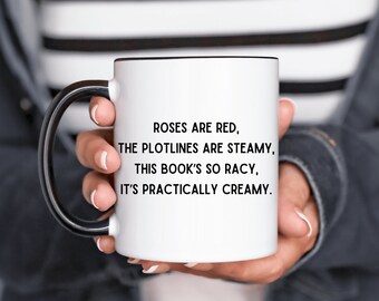 May include: White ceramic mug with a black handle and rim, featuring the text "ROSES ARE RED, THE PLOTLINES ARE STEAMY, THIS BOOK'S SO RACY, IT'S PRACTICALLY CREAMY." The mug is held by a person.