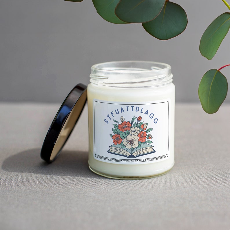 Może przedstawiać: Biały słoik ze świecą z czarną pokrywką. Słoik ma etykietę z kwiatowym wzorem i tekstem "STFUATTDLAGG". Etykieta zawiera r&oacute;wnież tekst "COCONUT CREAM | 100% NATURAL SOY WAX | 9 OZ | HANDCRAFTED WITH LOVE".