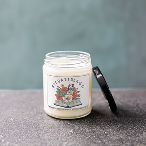 Może przedstawiać: Biały słoik ze świecą z czarną pokrywką. Słoik ma etykietę z tekstem "STFUATTDLAGG" i kwiatowy wz&oacute;r z książką pod kwiatami.