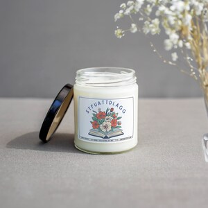 Może przedstawiać: Biała świeca z czarną pokrywką i etykietą z napisem "STFUATTDLAGG" z kwiatowym wzorem i książką.