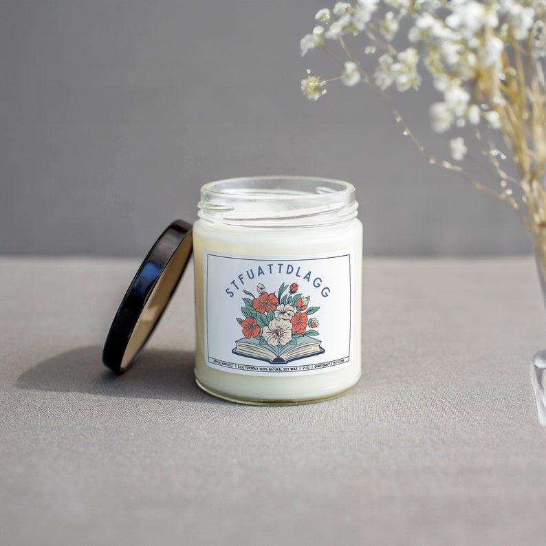 Może przedstawiać: Biała świeca z czarną pokrywką i etykietą z napisem "STFUATTDLAGG" z kwiatowym wzorem i książką.