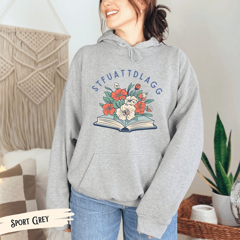 May include: A gray hooded sweatshirt with the text "STFUATTDLAGG" printed in blue on the front. The text is surrounded by a floral design with red, orange, and white flowers. The sweatshirt is shown on a model wearing blue jeans.