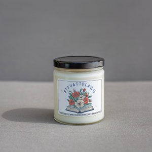 Może przedstawiać: Biały słoik ze świecą z czarną pokrywką i etykietą z napisem "STFUATTDLAGG" z kwiatowym wzorem i otwartą książką. Etykieta zawiera r&oacute;wnież napis "Eco-Friendly 100% Natural Soy Wax."