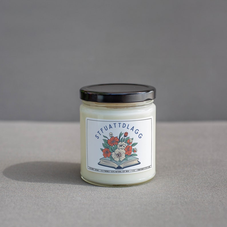 Może przedstawiać: Biały słoik ze świecą z czarną pokrywką i etykietą z napisem "STFUATTDLAGG" z kwiatowym wzorem i otwartą książką. Etykieta zawiera r&oacute;wnież napis "Eco-Friendly 100% Natural Soy Wax."