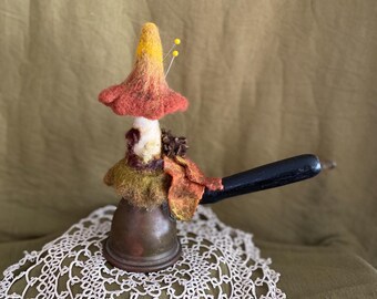 Puede incluir: Una figurita de hongo de fieltro caprichosa con un gorro amarillo y naranja, detalles de fieltro marrón y un mango de madera negro. La figurita está posada sobre una olla de cobre vintage y un encaje blanco.
