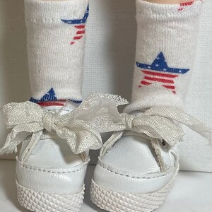 May include: White canvas shoes with white laces and a white bow. The shoes have a textured sole. The shoes are being worn by a doll with white socks with red and blue stars.