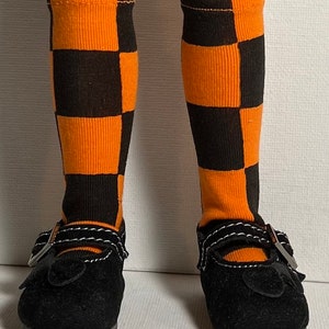 May include: A pair of knee-high socks featuring a black and orange checkered pattern, worn with black shoes that have a single strap across the top, a silver-toned buckle, and decorative white stitching along the edges of the strap.