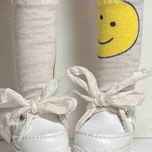 May include: A pair of white sneakers with white laces tied in bows. The sneakers are worn with white socks with a yellow smiley face design on one sock.