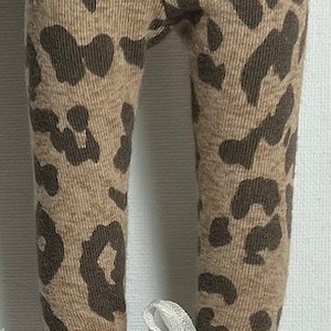 May include: A pair of brown and tan leggings with a leopard print pattern. The leggings are worn with white sneakers and a white ribbon tied around the ankles.