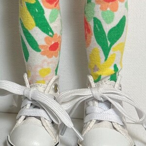 May include: A pair of white canvas high-top sneakers with white laces. The sneakers are worn by a doll with floral print knee-high socks.