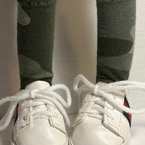 May include: A pair of white sneakers with red and green stripes on the sides. The sneakers are worn with green and white camouflage socks.