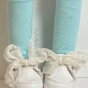 May include: A pair of white sneakers with white ribbon laces. The sneakers are worn by a doll with light blue socks.
