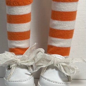 May include: A pair of white canvas shoes with orange and white striped socks. The shoes have white laces tied in a bow.
