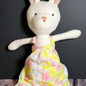 May include: A white plush bunny wearing a yellow and pink floral print overall. The bunny has long, floppy ears with pink fabric on the inside.